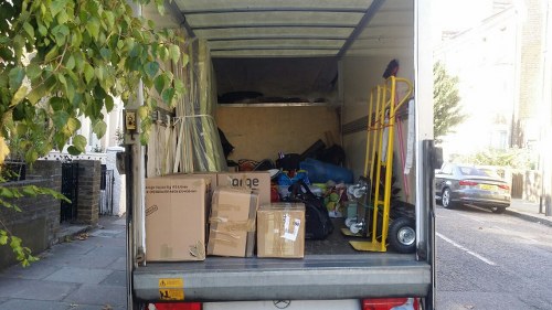 The image shows the rear of a small commercial vehicle parked on a street, with the back door open to reveal an interior filled with various types of rubbish. Inside, there are multiple cardboard boxes, some flattened and others still in their upright form, with clear print and labels visible on their surfaces. The boxes contain miscellaneous waste, possibly packaging materials, and are stacked towards the left side of the vehicle's cargo area. On the right, there is a yellow hand truck leaning against the vehicle wall, alongside a small black dolly or trolley and a few scattered plastic bags filled with waste. The environment around the vehicle suggests an urban area with pavement and parked cars visible along the street, and green foliage partially framing the scene on the left. The lighting indicates daytime, with natural sunlight illuminating the scene, emphasizing the textures of the cardboard, plastic, and metal surfaces. This setup is consistent with rubbish removal services in Lambeth, provided by Commercial Waste Lambeth, focused on clearing various waste and rubbish efficiently from commercial properties or urban locations.