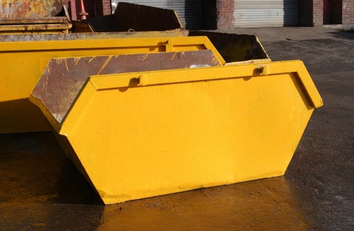 A large, rectangular yellow skip with a smooth, painted metal surface positioned on a concrete ground outdoors. The skip features reinforced edges and a flat top, showing some signs of use such as minor scratches and dirt. It is situated in an industrial or commercial outdoor area, with other similar skips visible partially in the background. The lighting indicates daytime, with natural light casting soft shadows on the ground and highlight on the skip’s glossy finish. This image is relevant to rubbish and waste removal services provided by Commercial Waste Lambeth, emphasizing their capacity to handle large-scale waste collection and disposal tasks in the Lambeth area. The surroundings include a paved driveway or yard typical of waste management sites, with some brickwork and metal structures faintly visible behind the skips, consistent with industrial or commercial waste management environments.