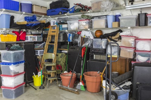 A storage area filled with various stored items including clear plastic storage boxes with red, black, and blue lids stacked on shelves and on the floor, a wooden ladder leaning against the shelving unit, a yellow plastic bucket, several plastic plant pots, a green bag, brooms, and a mop placed in front of a wall of shelving units containing tools, boxes, sports equipment, and miscellaneous household items. The environment appears to be a garage or storage room with a concrete floor, and the items are organized in a manner typical for waste removal or clearance services provided by Commercial Waste Lambeth in the Lambeth area, South London.