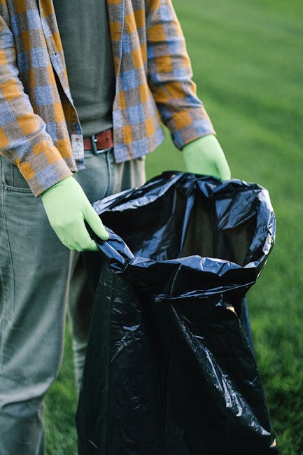 A person wearing a yellow and gray checkered shirt, green gloves, and a dark green t-shirt stands outdoors on a grassy area, holding open a large black rubbish bag lined with a shiny black plastic int
