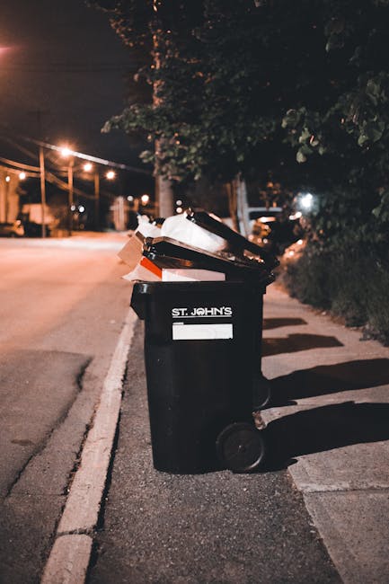 A large, weathered green skip filled with a mixture of cardboard boxes, paper packaging, and miscellaneous waste materials is situated outdoors on a paved surface, with additional cardboard and packin